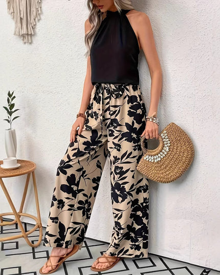 Woman wearing a black halter top and floral pants, holding a straw bag, standing against a white wall.