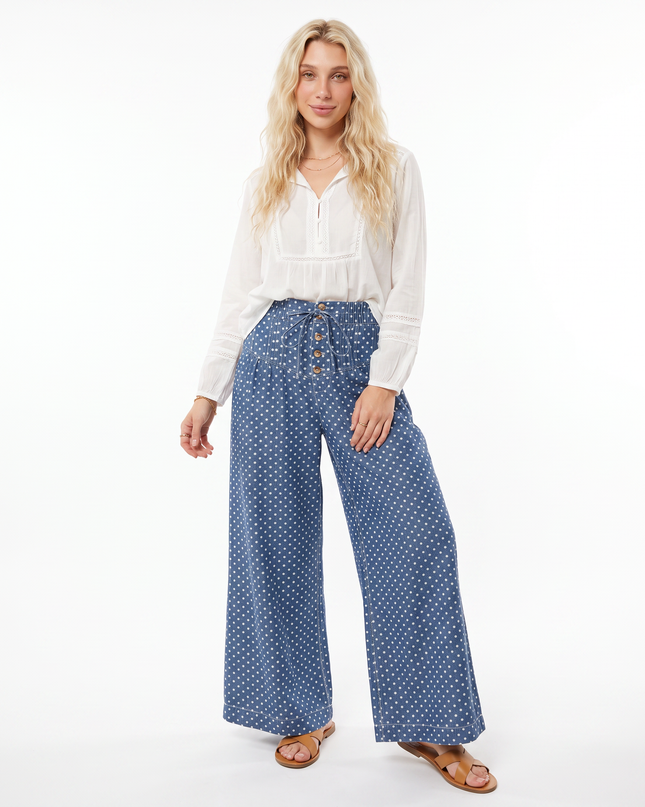 A woman modeling a pair of wide-leg blue pants with a white polka dot pattern, paired with a white blouse and brown sandals, against a plain white studio background.