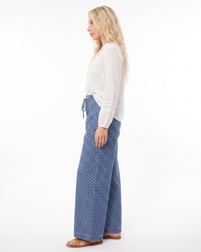 A woman stands in profile wearing a white long-sleeve top and blue wide-leg pants with a small white polka dot pattern, photographed against a plain white studio background.
