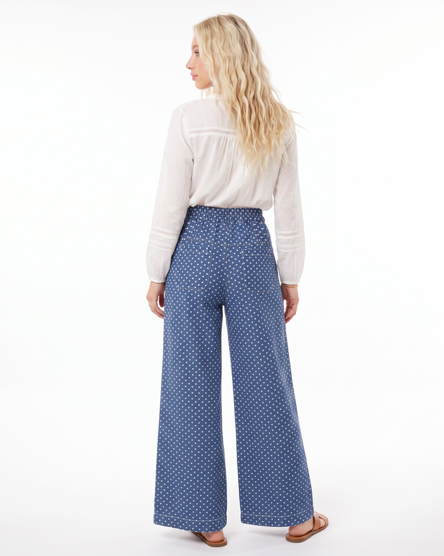 A woman shown from the back wearing high-waisted, wide-leg blue pants with a small white polka dot pattern, paired with a white blouse and brown sandals, against a plain white background.