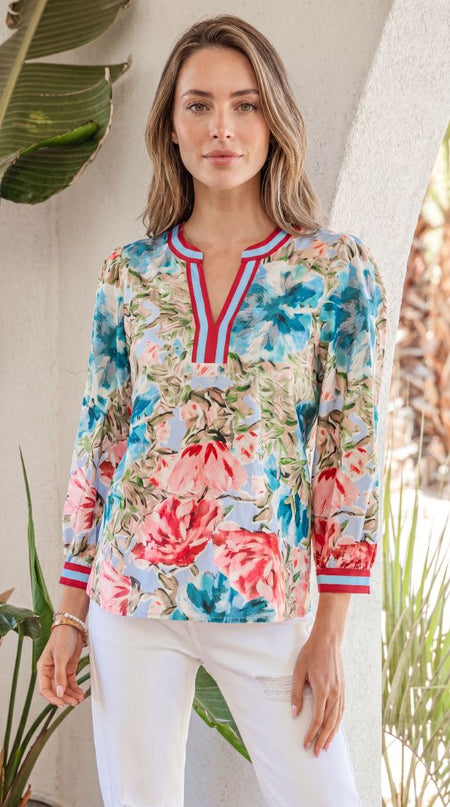 Woman wearing a colorful floral long-sleeve top with white pants, standing against a neutral background with plants.