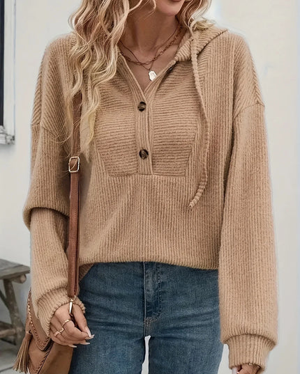 Person wearing a beige ribbed sweater with a hood, holding a brown bag indoors.