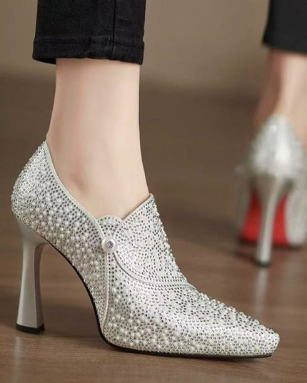 Silver high-heeled shoes with pearl and rhinestone details on a wooden floor.