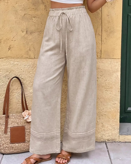 Person wearing beige wide-leg pants standing next to a textured wall and straw bag.