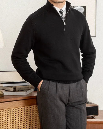 Man wearing a black quarter-zip sweater and gray pants in a room with a lamp and books on a table.