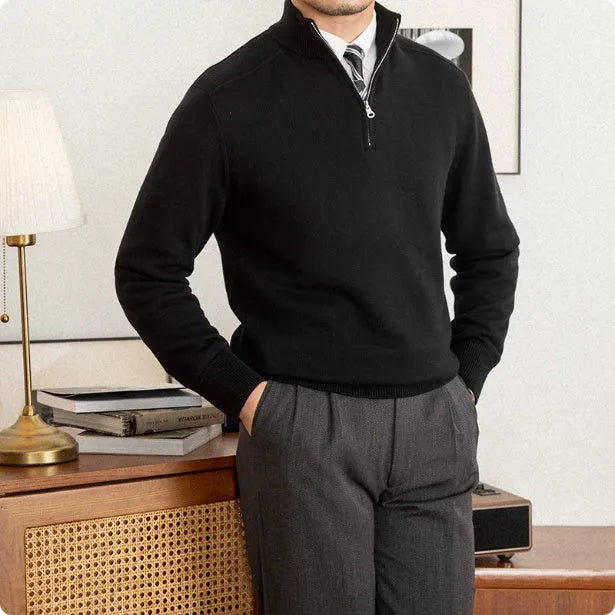 Man wearing a black quarter-zip sweater and gray pants in a room with a lamp and books on a table.