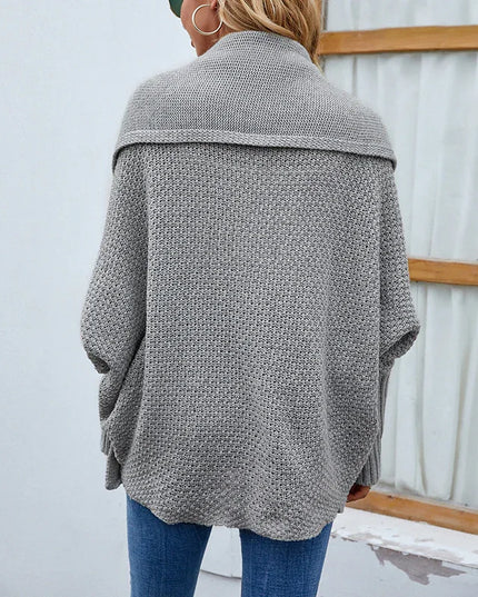 Person wearing a gray knitted cardigan and blue jeans against a white wall.