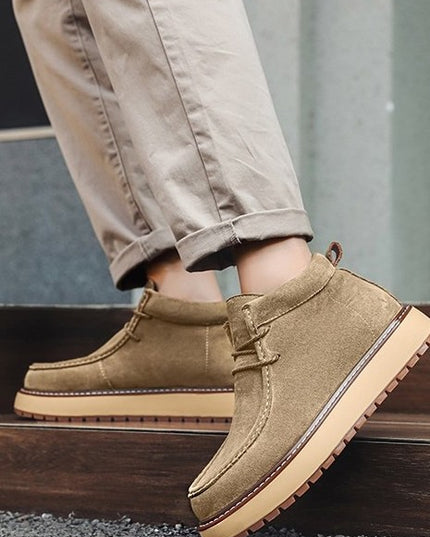 Tan suede shoes with brown soles worn by a person in beige pants.