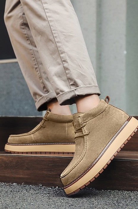 Tan suede shoes with brown soles worn by a person in beige pants.