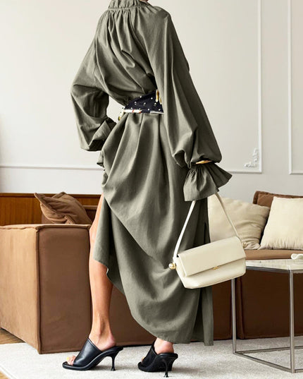 Person wearing a green dress with long sleeves and a belt, holding a cream handbag in a living room.