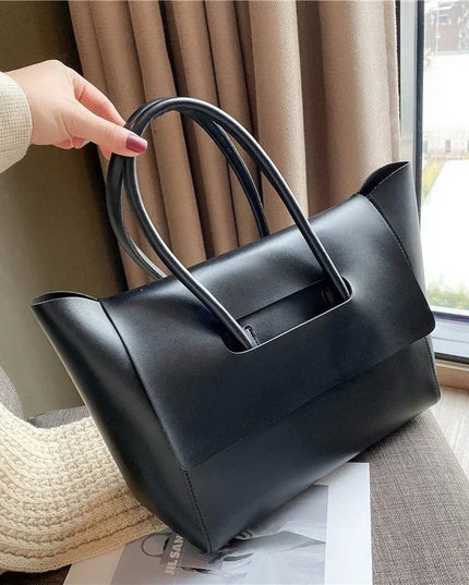 Black handbag held by a person in a home setting with curtains and a window in the background.