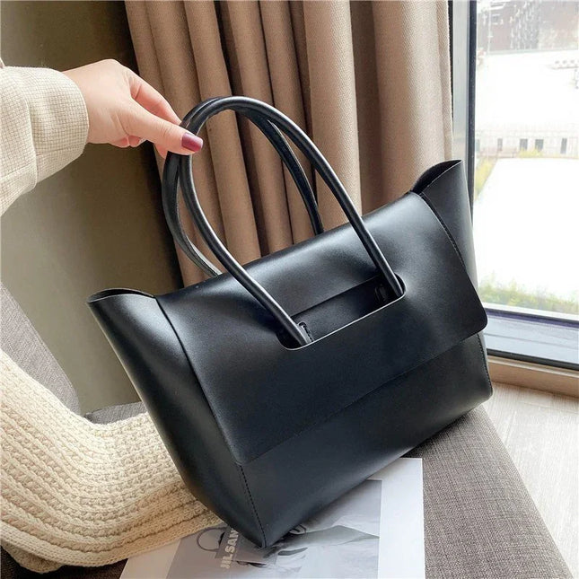 Black handbag held by a person in a home setting with curtains and a window in the background.
