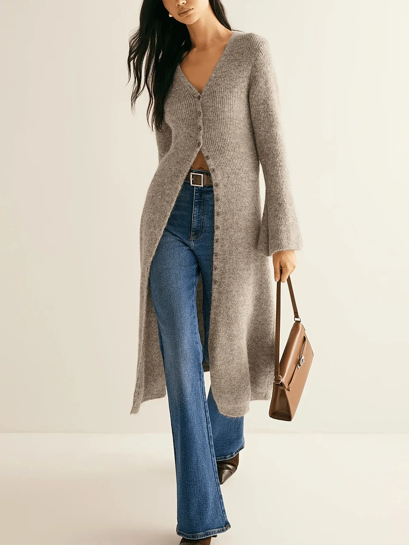 Woman wearing a long gray cardigan, blue jeans, and holding a brown handbag on a beige background