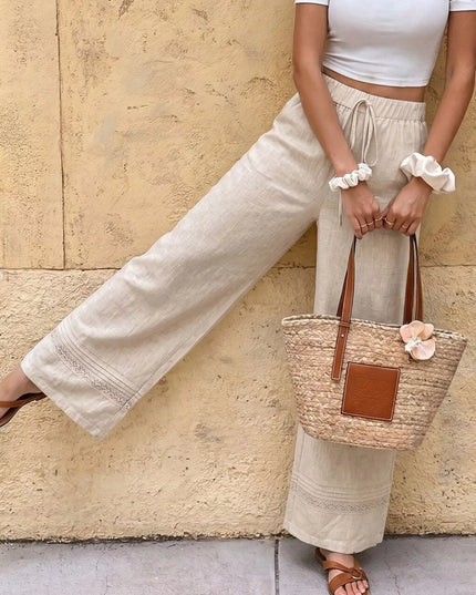 Person wearing light-colored pants and a white top, holding a straw bag against a beige wall.