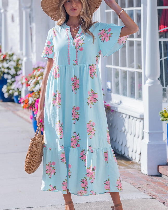 Woman wearing a light blue floral dress with a straw hat and bag, standing on a sidewalk.