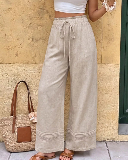 Person wearing beige wide-leg pants with a white top and brown sandals, standing against a stone wall.