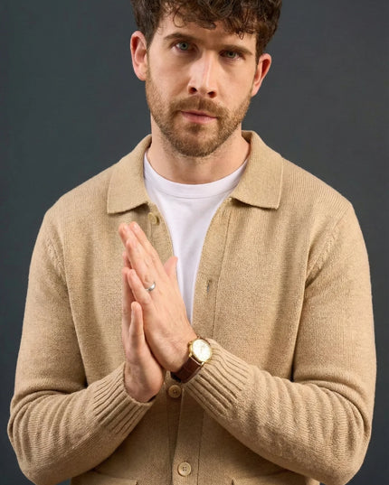 Man wearing a beige jacket over a white shirt with hands clasped together against a dark background