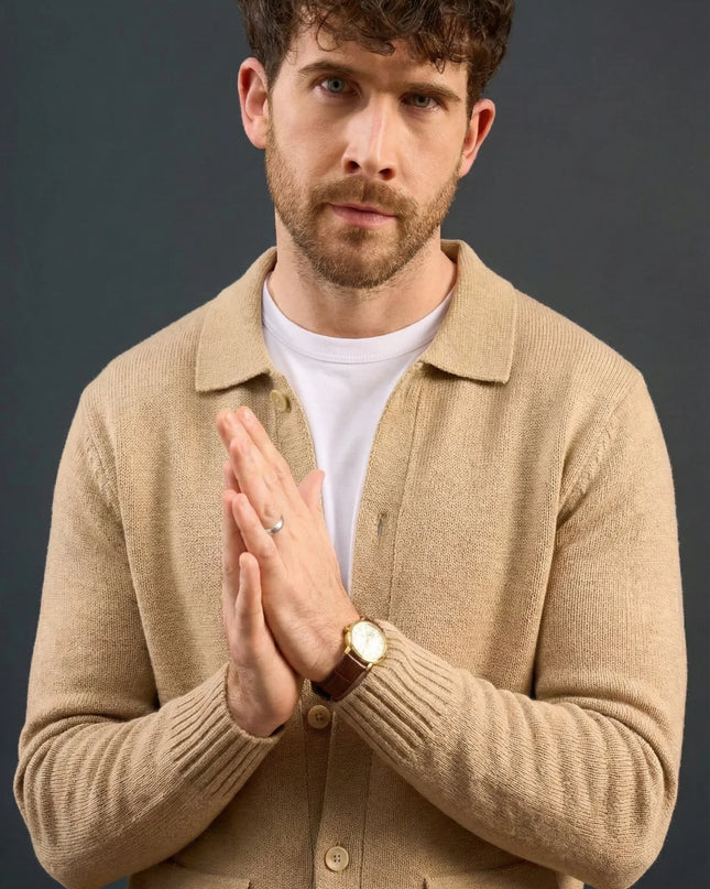 Man wearing a beige jacket over a white shirt with hands clasped together against a dark background