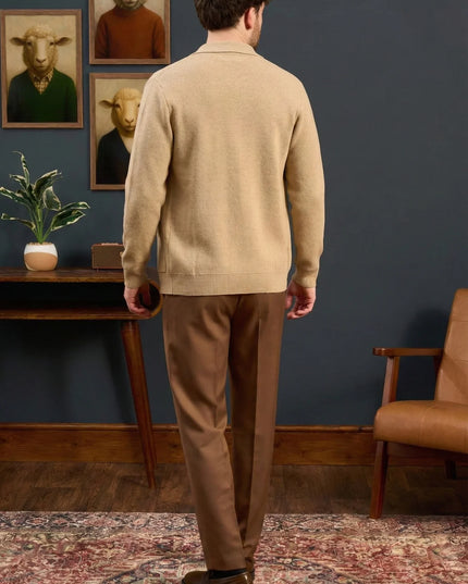 Man in beige sweater and brown pants standing in a room with blue walls, a wooden table, and a chair.