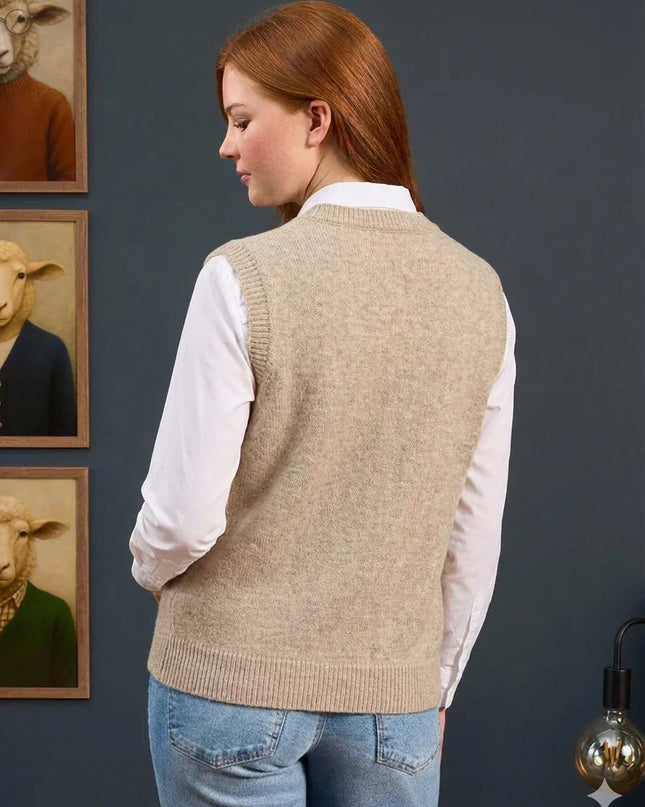 Woman wearing a beige knitted vest over a white shirt with a gray wall and framed pictures of animals in the background.
