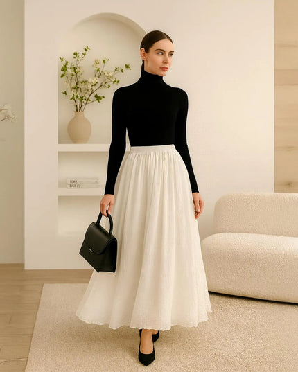 Woman in black turtleneck and white skirt holding a black handbag in a modern interior setting.