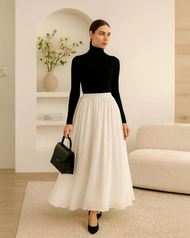 Woman in black turtleneck and white skirt holding a black handbag in a modern interior setting.