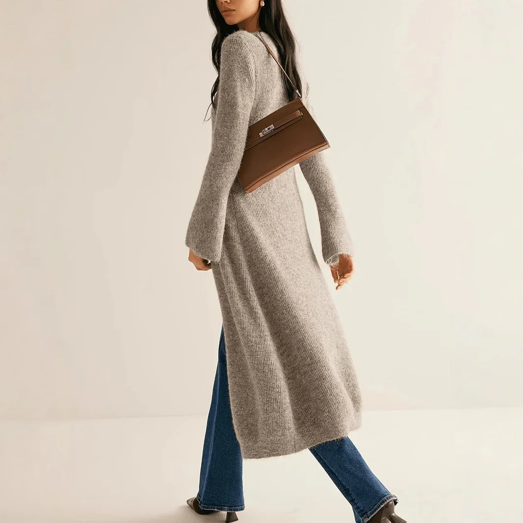 Woman wearing a long gray cardigan and blue jeans, holding a brown handbag on a beige background