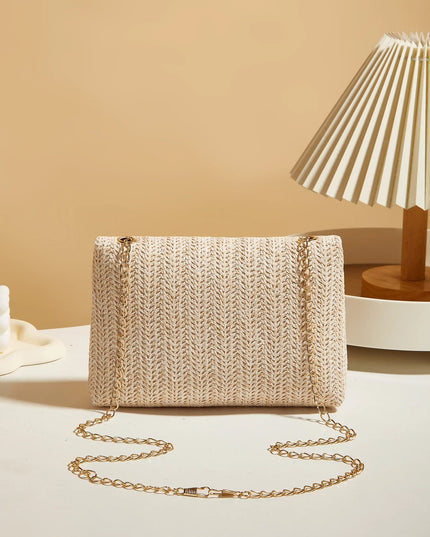 Beige woven handbag with a gold chain on a light surface with a beige wall and lamp in the background.
