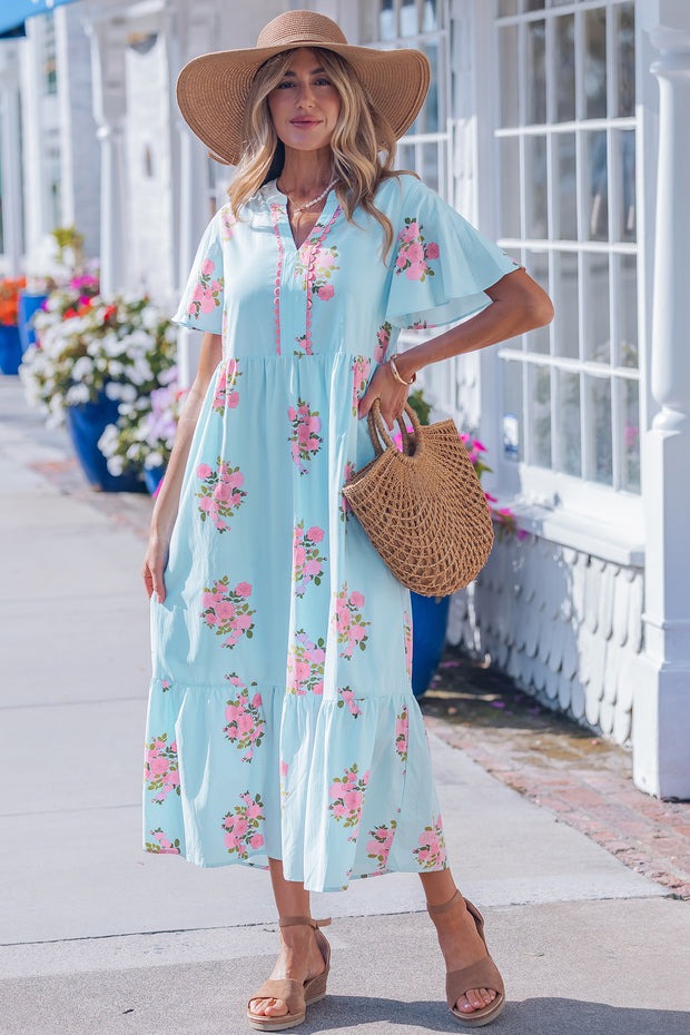 Woman in a floral dress with a straw hat and bag, standing on a sidewalk.