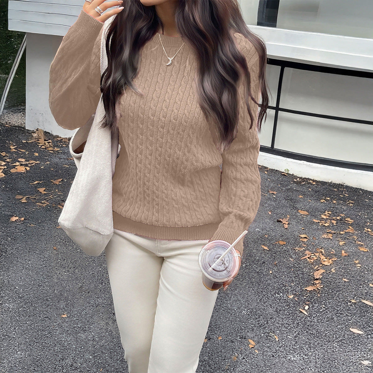 Person wearing a brown sweater and white pants, holding a drink, standing on a road with a building in the background.