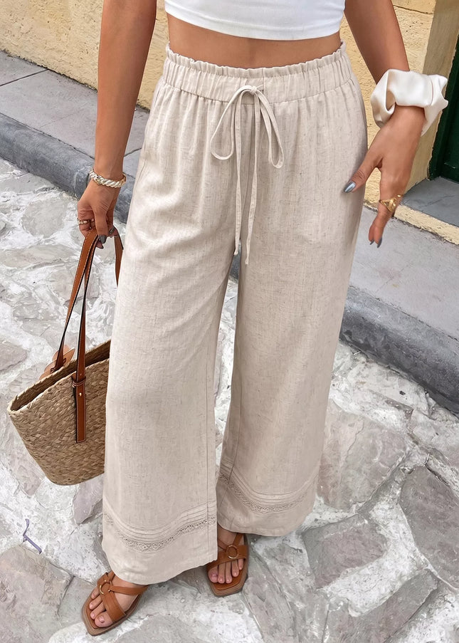 Person wearing beige wide-leg pants and a white top, holding a straw bag on a stone pavement.