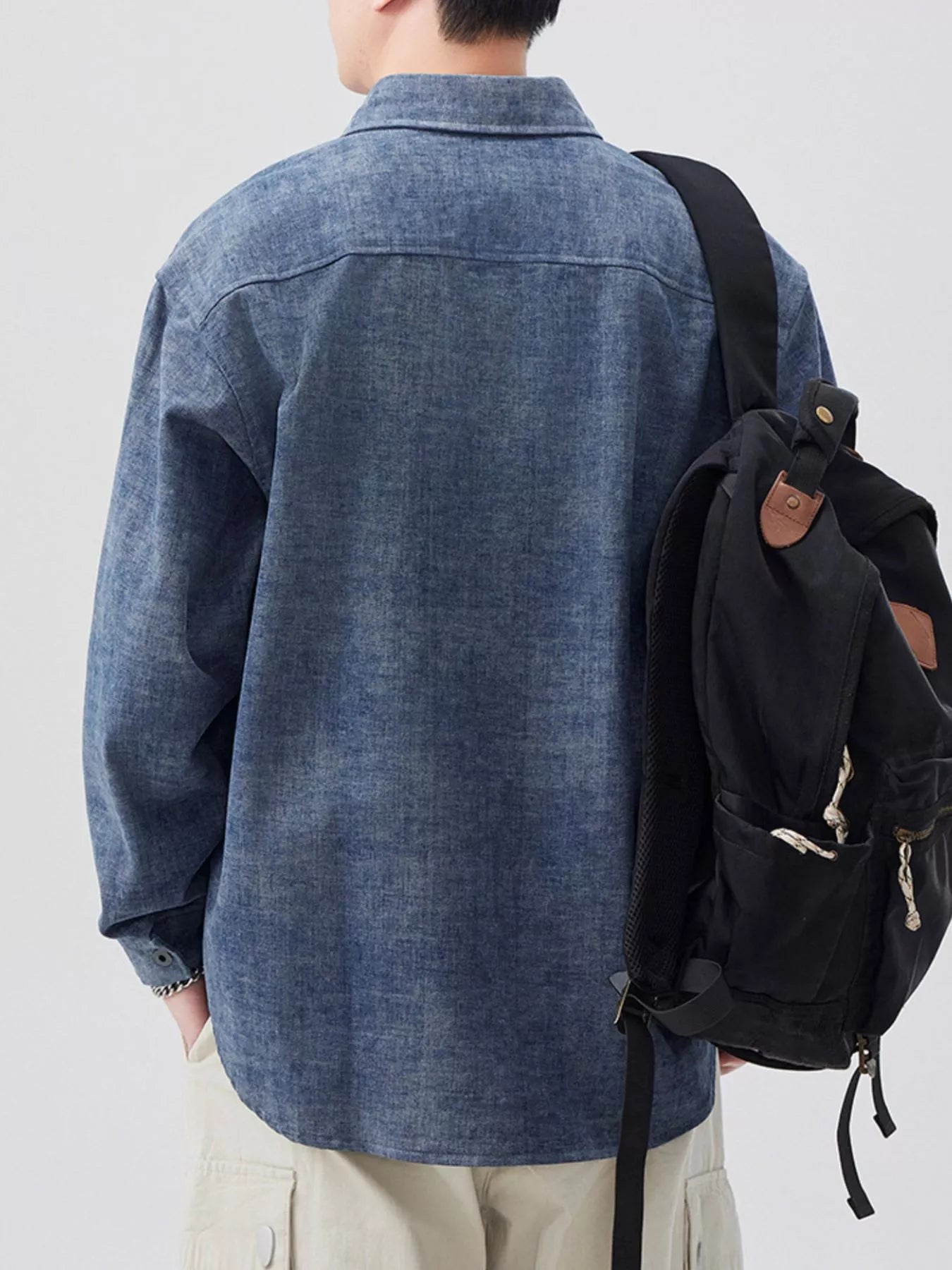 Person wearing a blue denim shirt and carrying a black backpack on a plain background