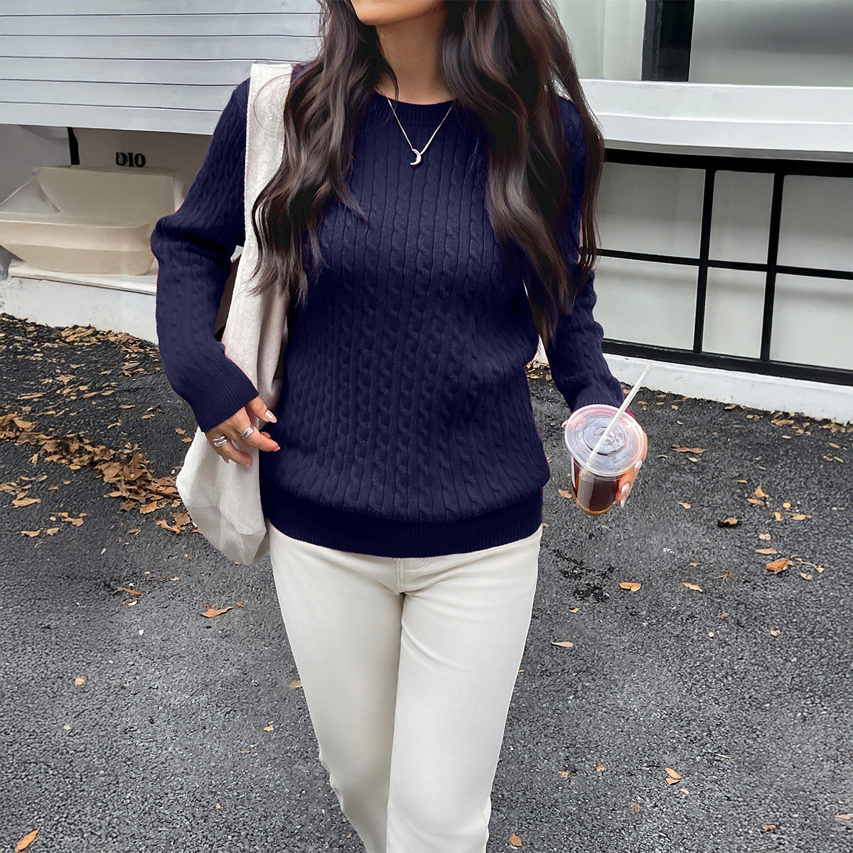 Woman wearing a navy blue sweater and white pants, holding a drink, standing on a street.