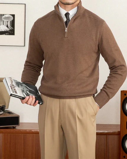 Man wearing a brown sweater and beige pants holding a book in a room with a record player and speakers.