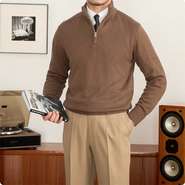 Man wearing a brown sweater and beige pants holding a book in a room with a record player and speakers.