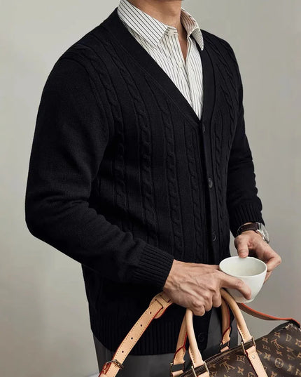 Person wearing a dark cable knit cardigan holding a cup and a patterned bag against a neutral background