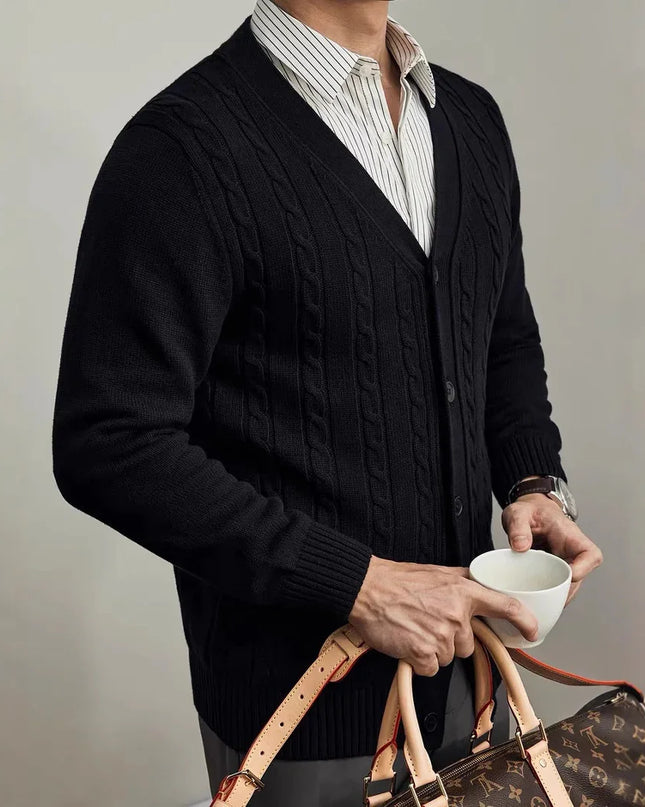 Person wearing a dark cable knit cardigan holding a cup and a patterned bag against a neutral background