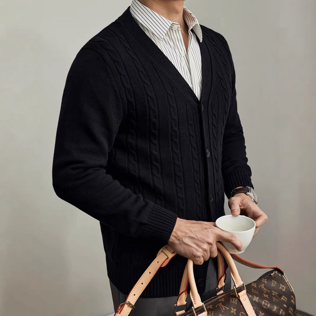 Person wearing a dark cable knit cardigan holding a cup and a patterned bag against a neutral background
