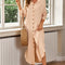 Woman in a light pink dress standing by a window holding a straw bag.